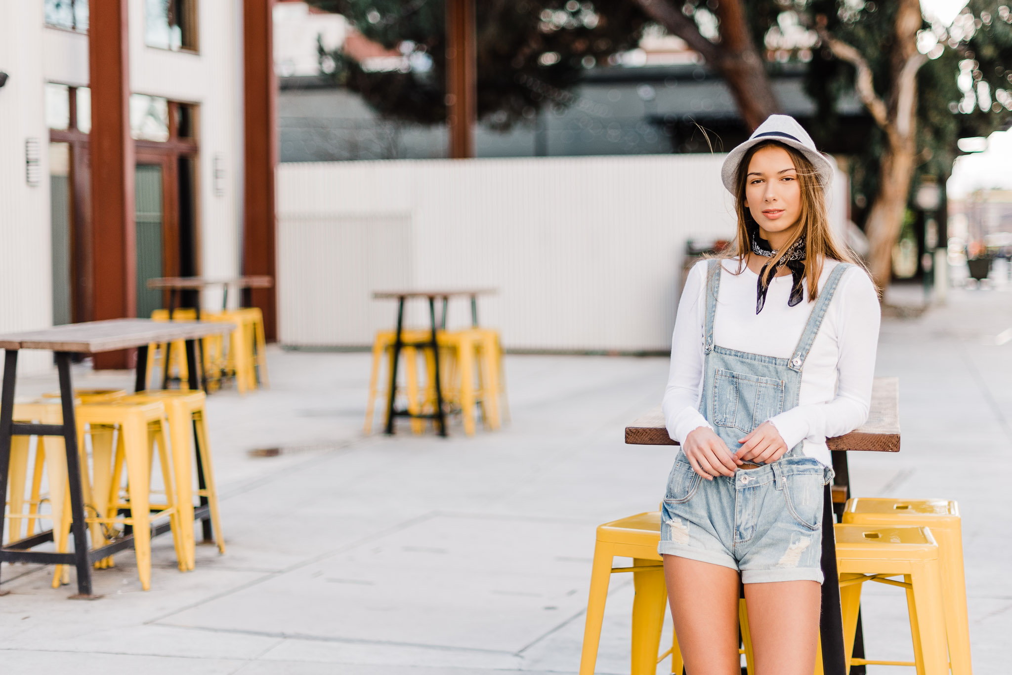 Teen wearing overalls in an urban setting with yellow chairs, by Emily Kim Photography.