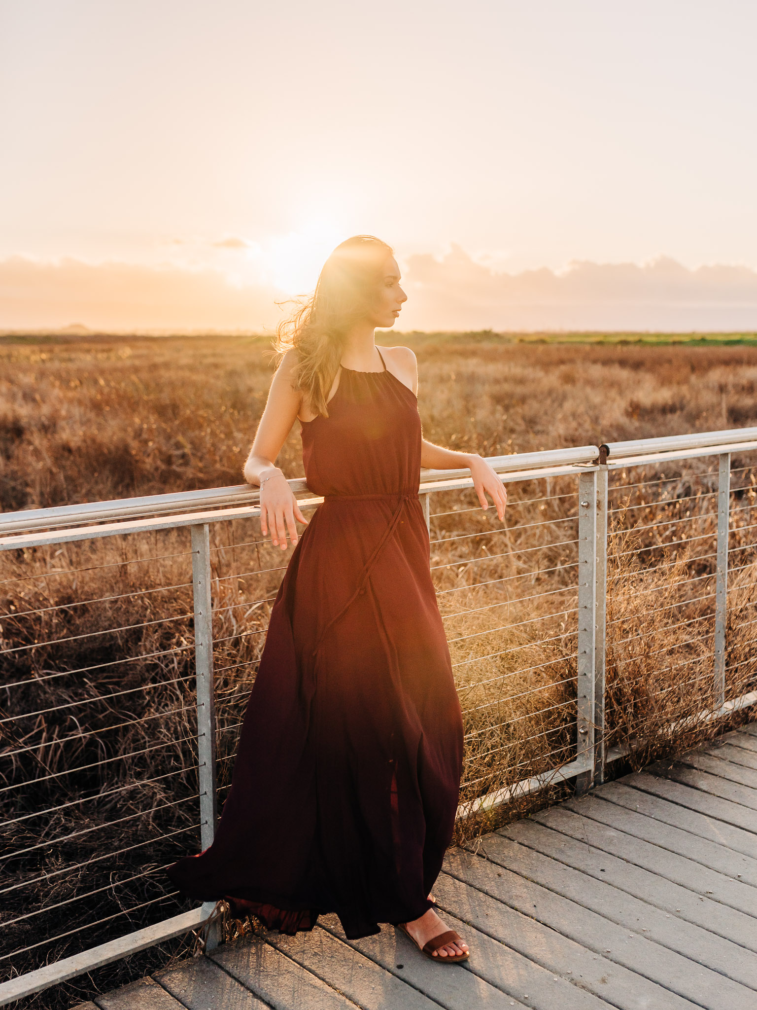 Teen in maroon dress leaning on railing with a sunset nature backdrop, by Emily Kim Photography.