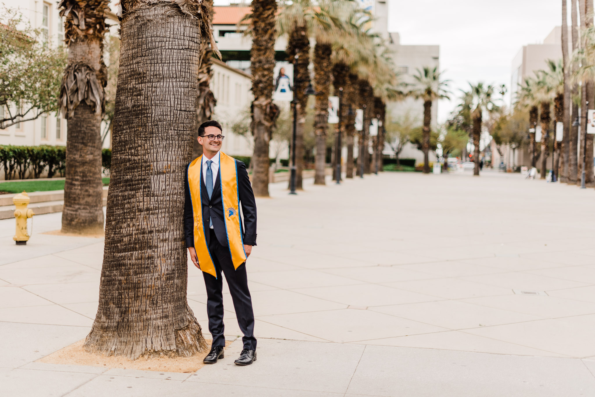 College grad leaning against palm tree, wearing a suite and stole.