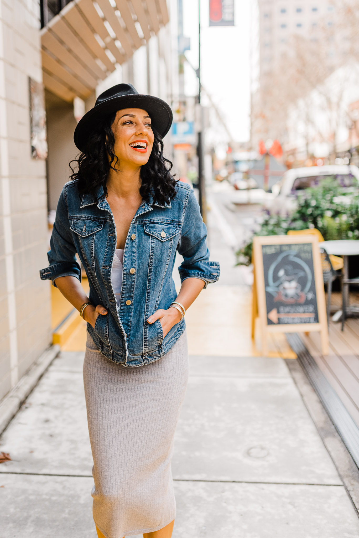 Woman walking down the sidewalk, looking away from the camera and laughing. She's wearing a gray maxi dress, denim jacket, and a black hat.