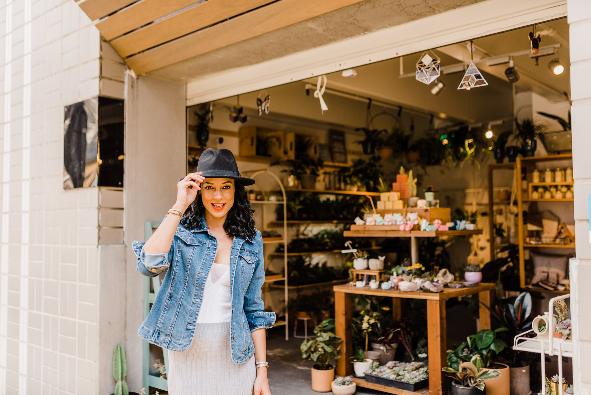Woman posing in front of a plant store, looking away from the camera and laughing. She's wearing a gray maxi dress, denim jacket, and a black hat.