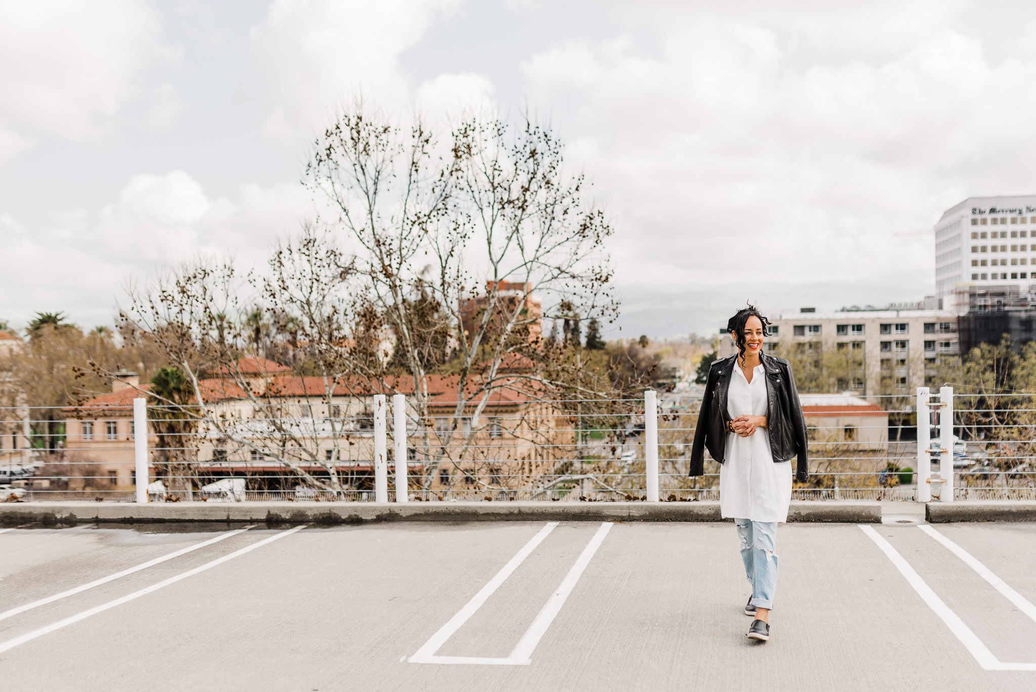 Fun portrait of a woman wearing a white tunic, black leather jacket, and light wash jeans. She's on top of a parking structure with San Jose's skyline in the background.