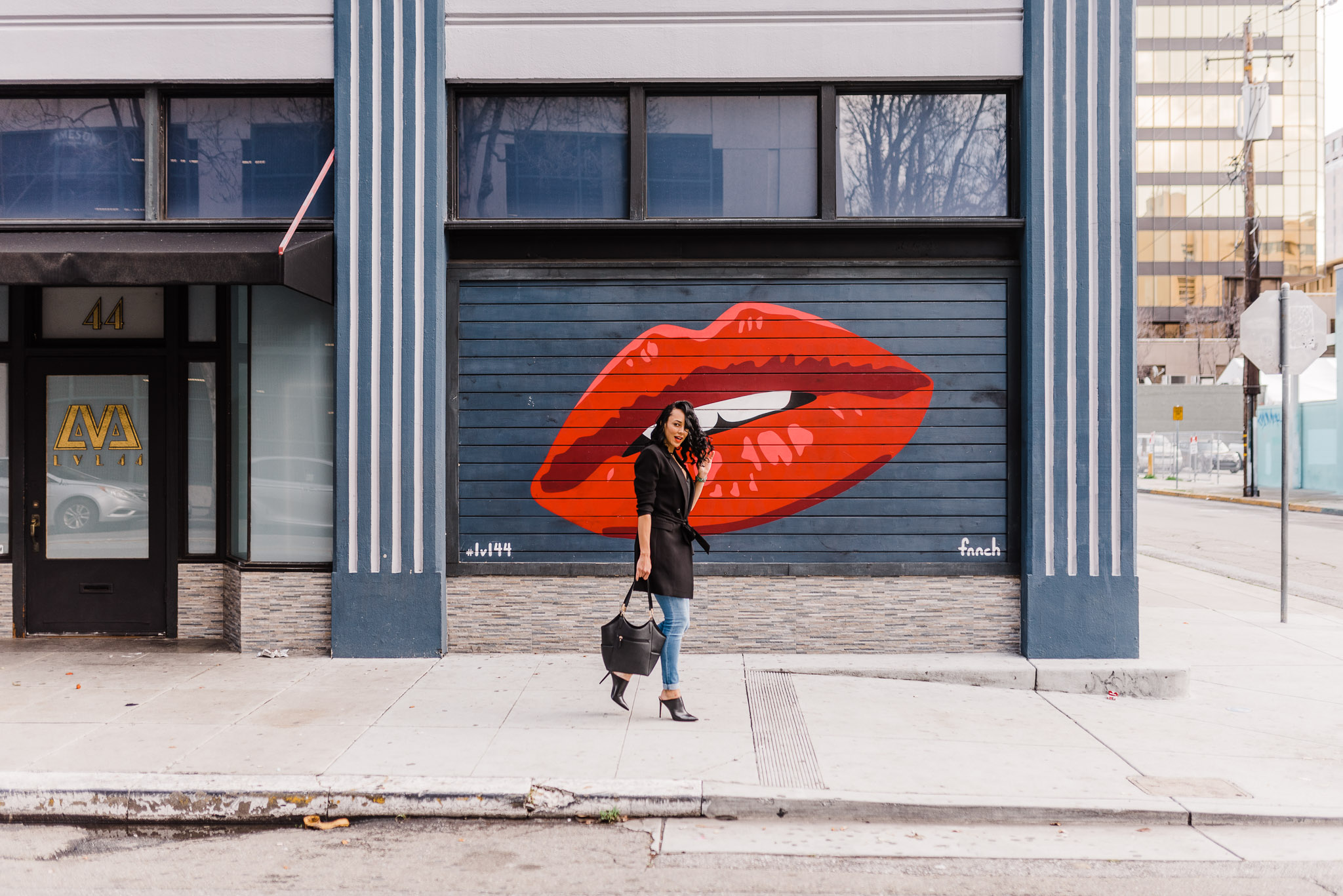 Woman walking down sidewalk in front of a red lipstick mural. She's wearing a black shirt, black blazer, and jeans.