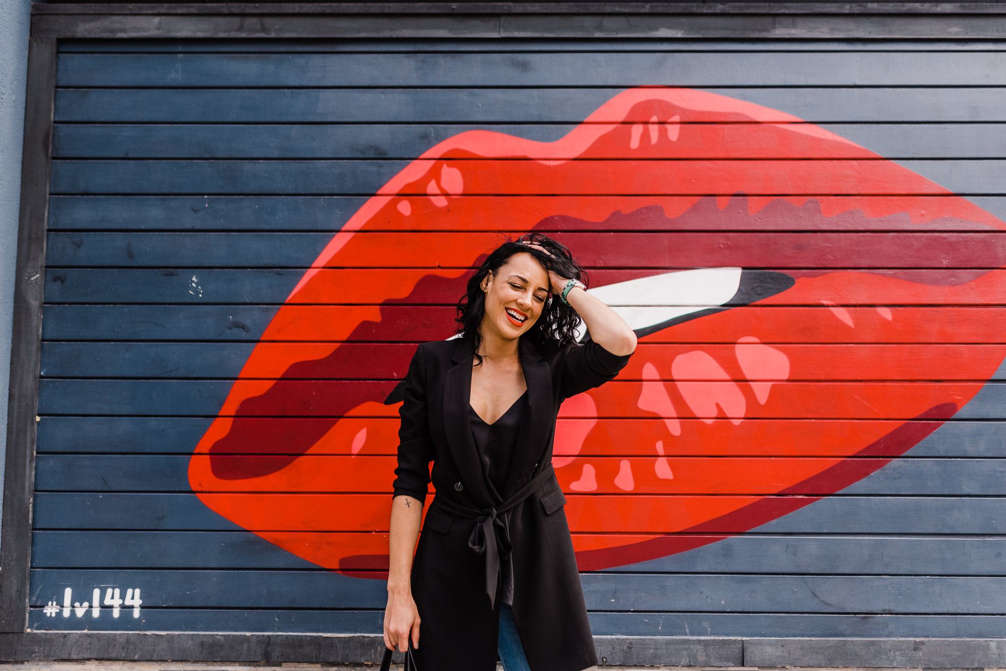 Woman posing in front of a red lipstick mural. She's wearing a black shirt, black blazer, and jeans.