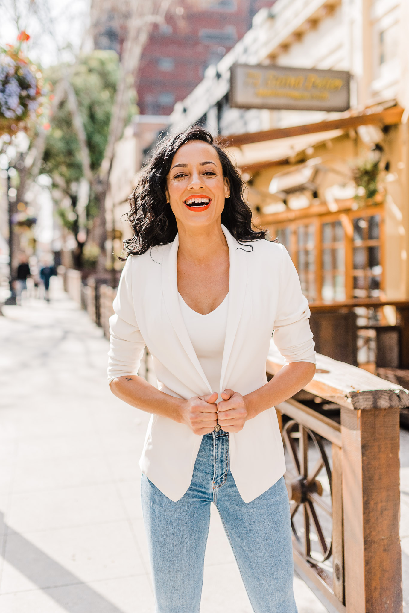 Woman wearing a white shirt, white blazer, and light wash jeans. She's holding her jacket and laughing.