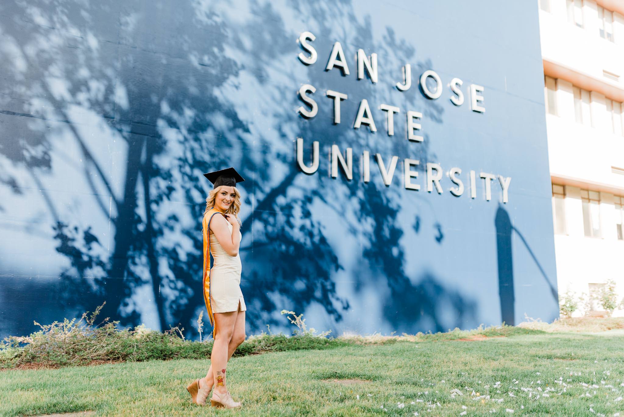 Grad holding stole and wearing graduation cap standing in front of San Jose State University sign