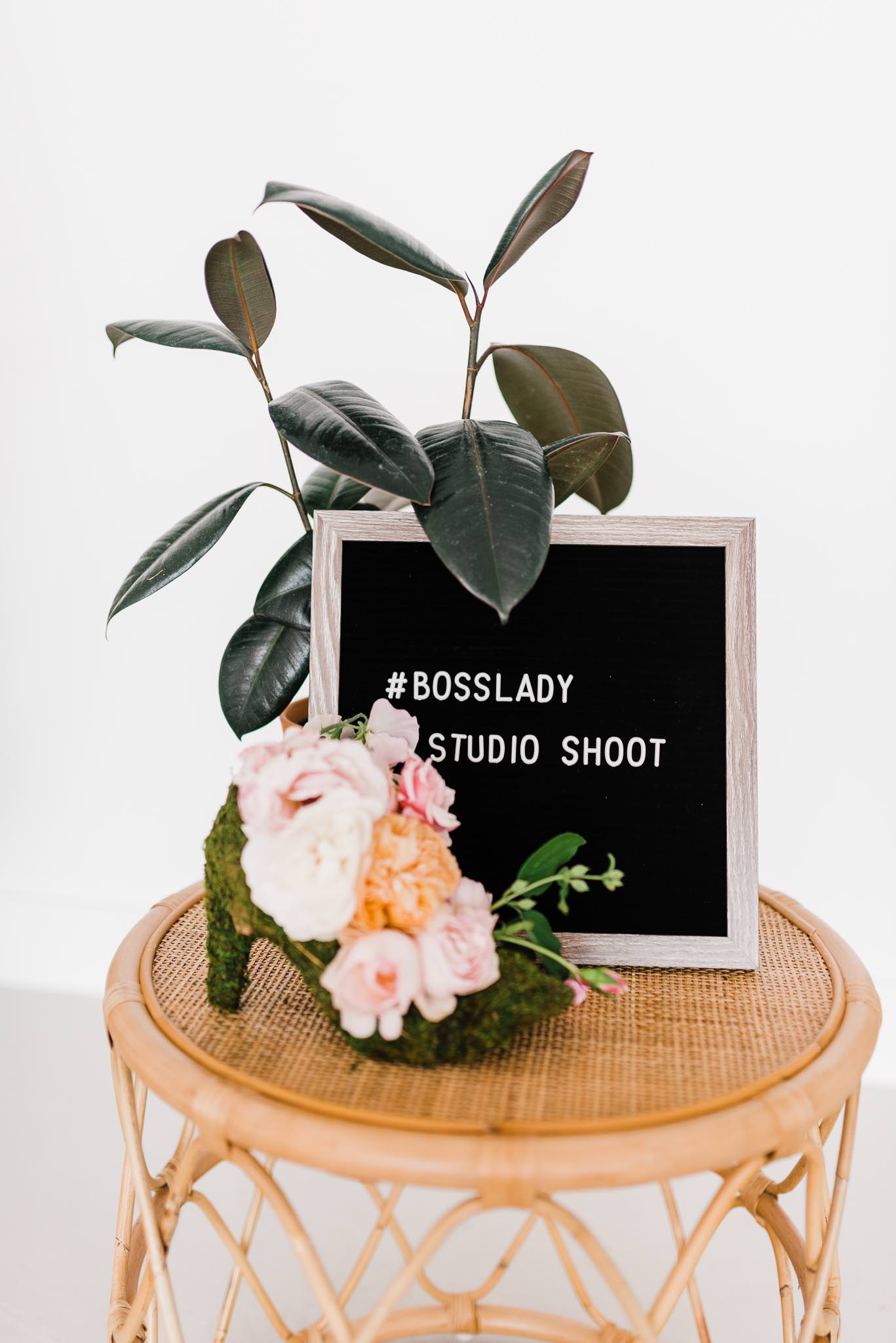 Florals in a moss high heel, with a black letter sign that says "#BossLady Studio Shoot."