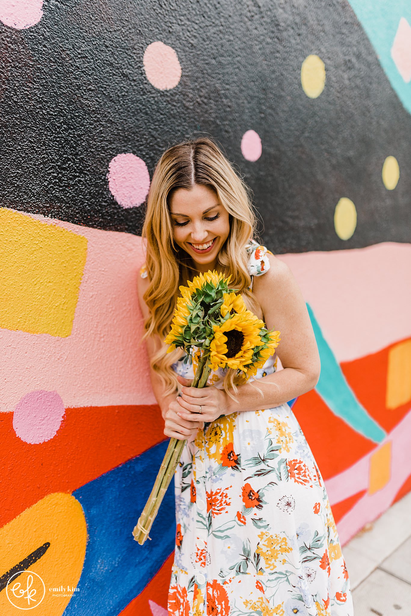 A woman leaning against a colorful mural holding sunflowers in San Jose.