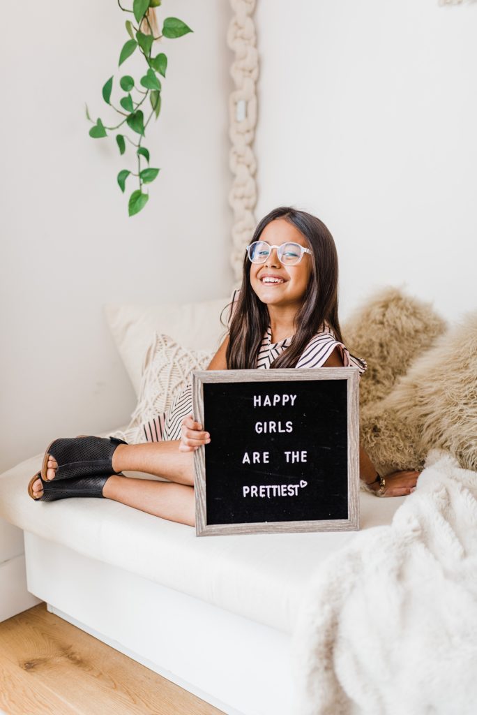 A girl wearing glasses holding a sign that says "Happy girls are the prettiest."