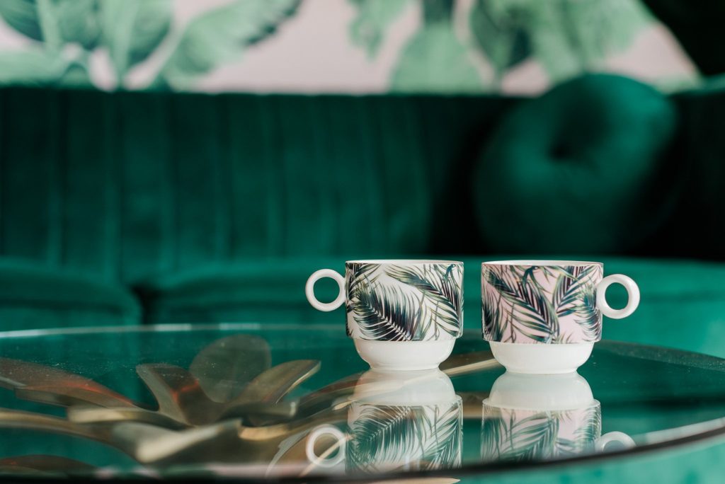 Two mugs with tropical print sitting on a glass table. One is pink and one is white. The backdrop is a velvet green couch.