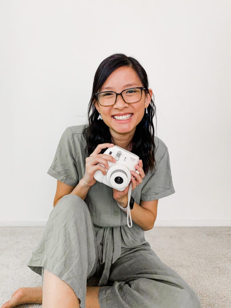 Woman wearing a green jumpsuit, sitting on the floor holding a fujifilm Instax camera.