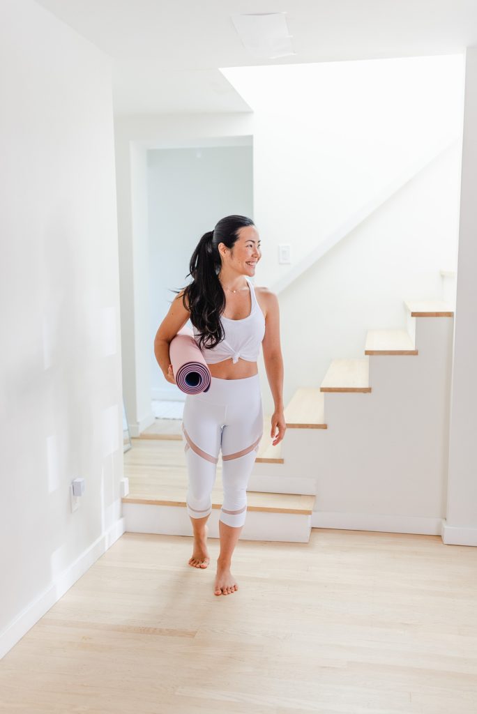 Woman in white workout clothes walking while carrying a rolled yoga mat.