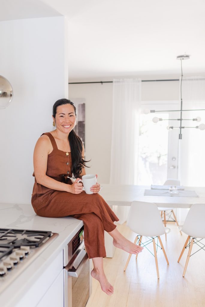 Woman in brown rompers sitting down on a kitchen counter while holding a white mug.
