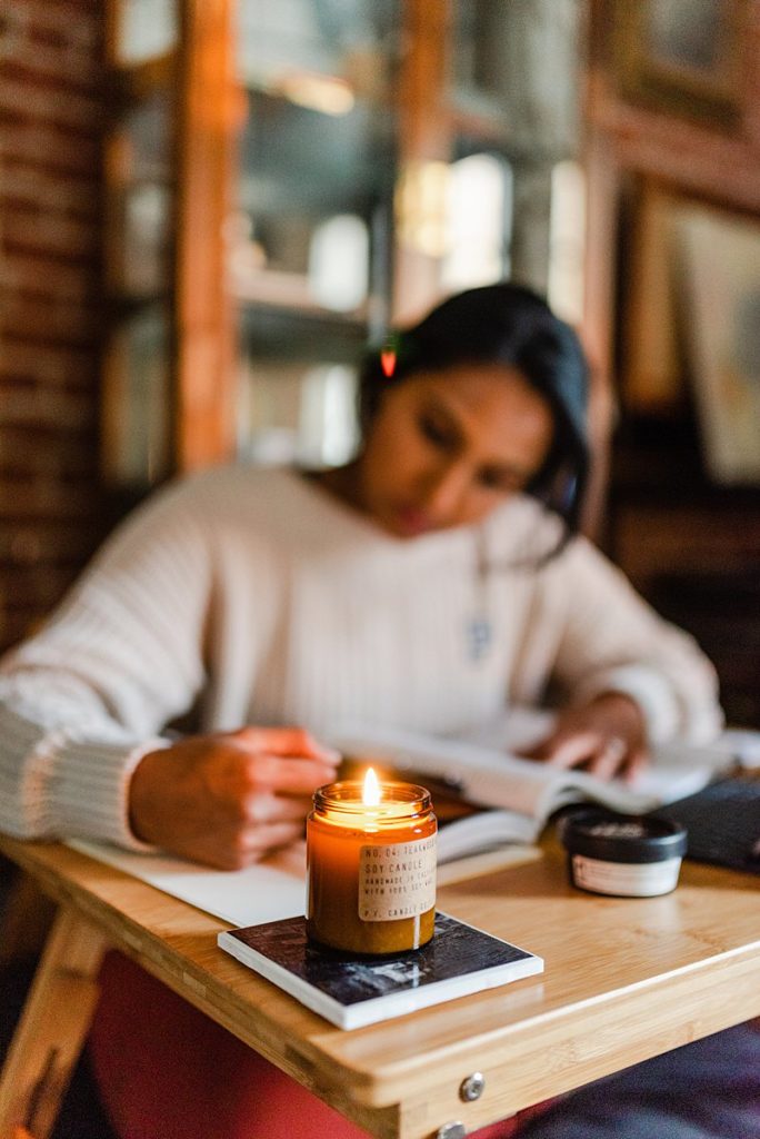 woman wearing a white sweater is writing something on an open book that is partly obscured by a lit candle. everything is out of focus except for the candle