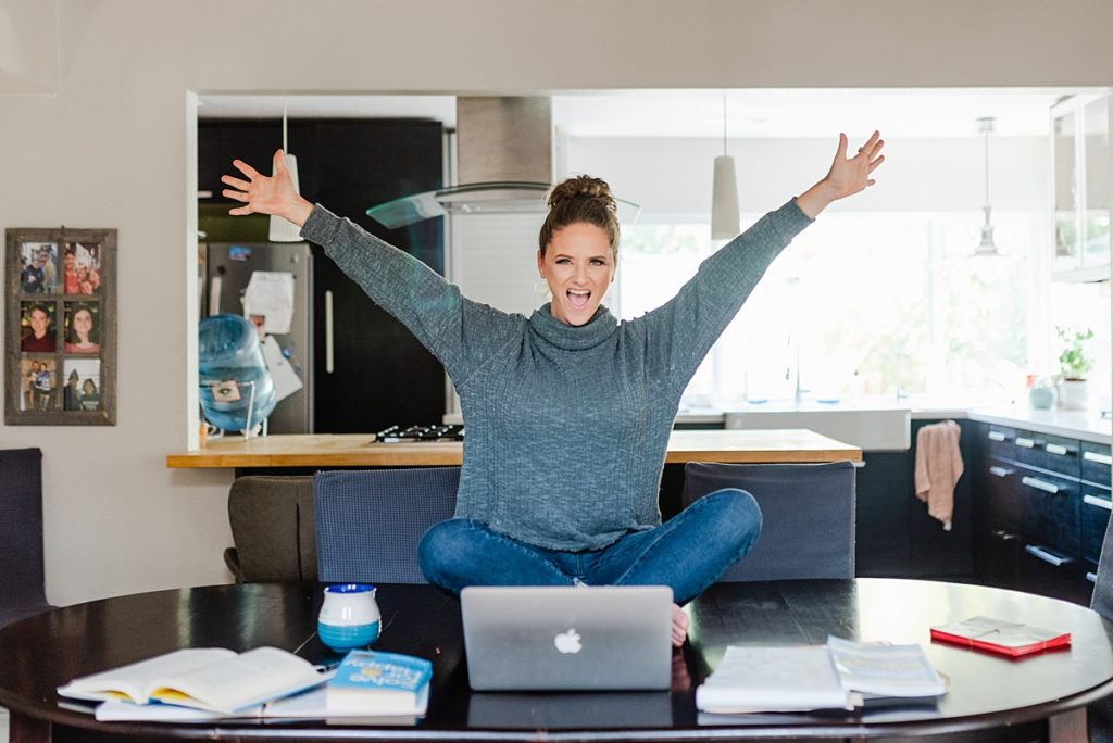 woman sitting cross-legged on top of a table raising both her arms in celebration with a laptop in front of her