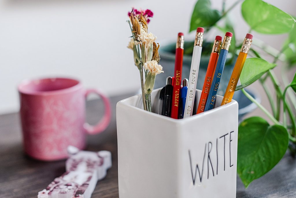 white pen holder with a couple of different colored pens and pencils that has the word "WRITE" on one side. there's a pink mug in the background that's out of focus.
