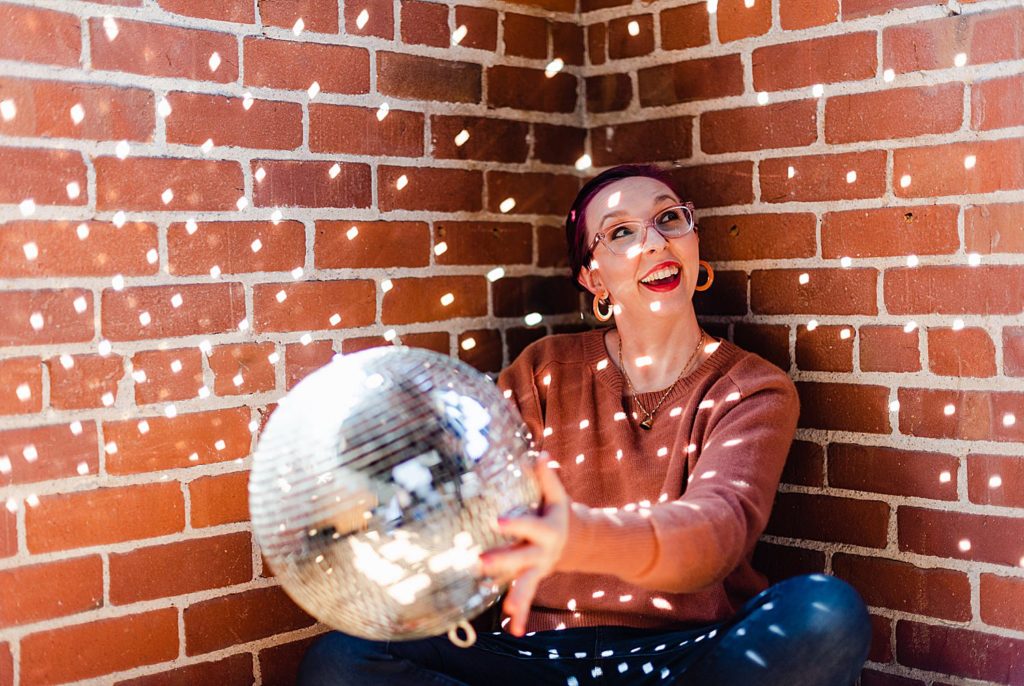 woman in brown sweater is holding a disco ball while sitting legs crossed on a corner. light is hitting the disco ball causing specks of light to scatter around her