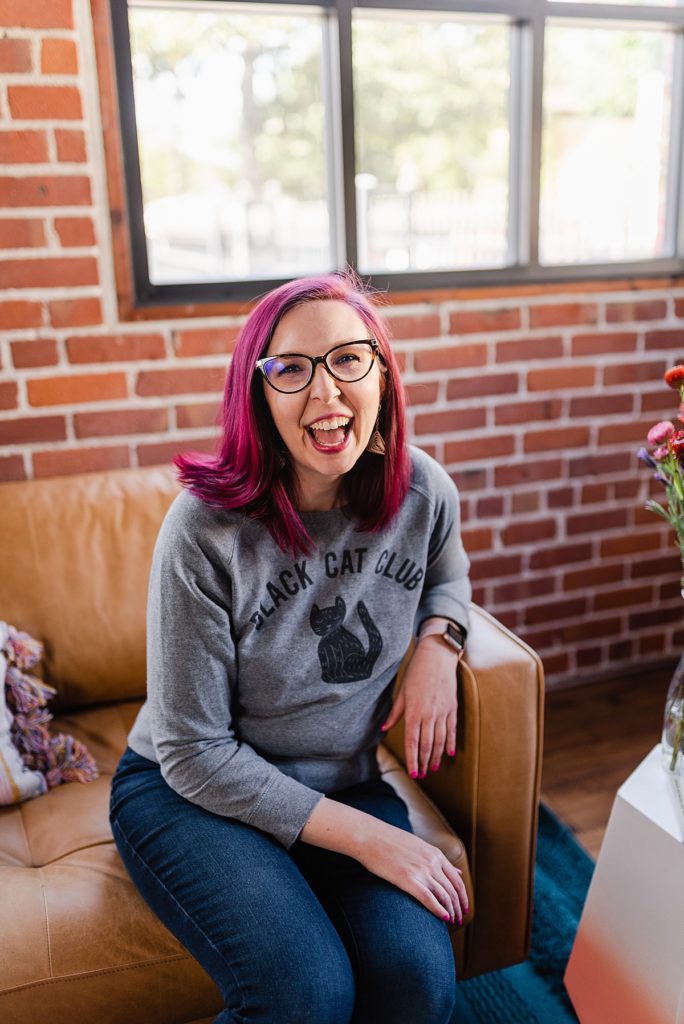woman with pink hair is smiling to the camera, she's wearing glasses, a gray sweater, and jeans