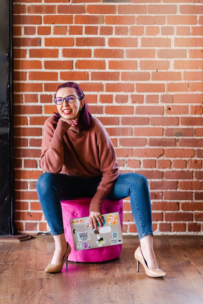 a woman is sitting on a pink ottoman with her chin on her hand and her elbow on her knee. she's holding her laptop on her other hand.