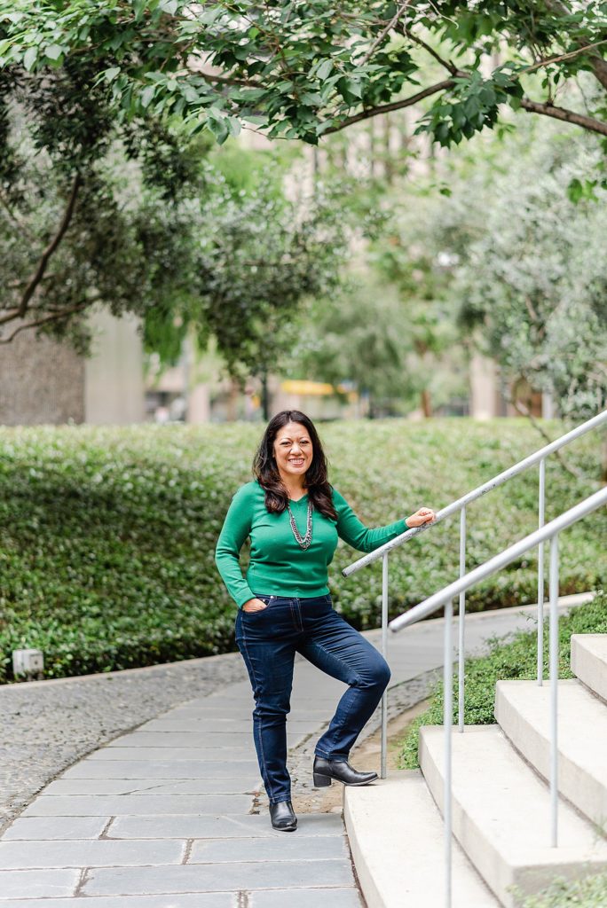 woman in green long-sleeved shirt is posing beside a stairway. she is holding the rails with one hand while her other hand is in her pocket, her foot is also on the first step