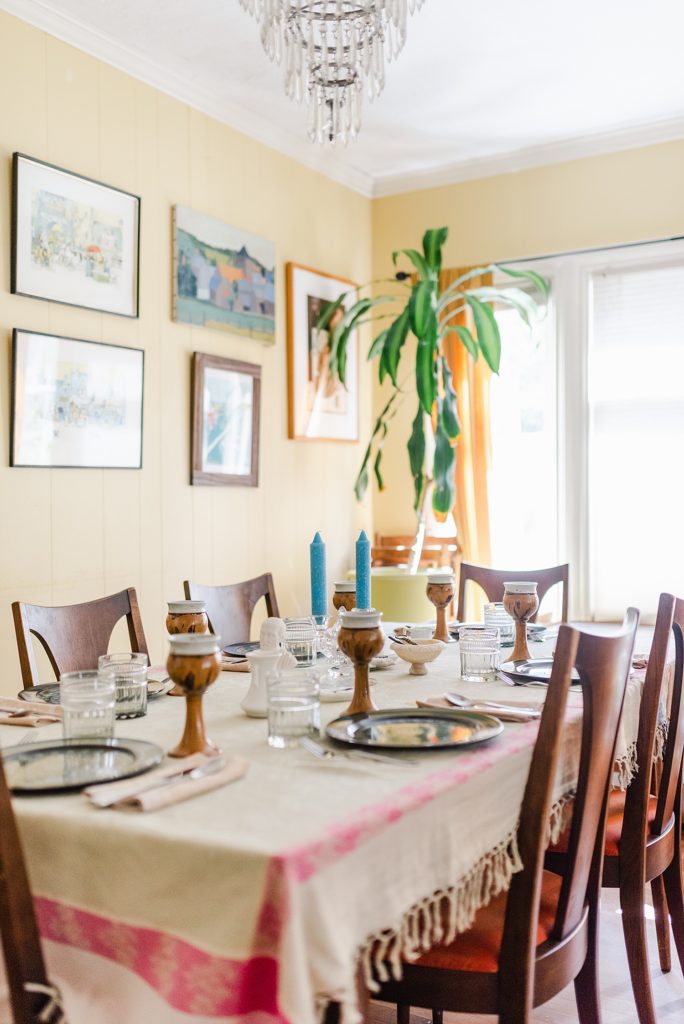 dining room with pastel yellow walls, a big plant in the corner beside the widow, wall arts, and crystal chandelier. the table is arranged with dark colored plates, two blue candles, glasses, tall ceramic glasses in gold, and cutlery on top of napkins.