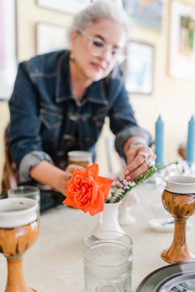 woman in the background is arranging a flower vase with one bright orange flower and some greenery