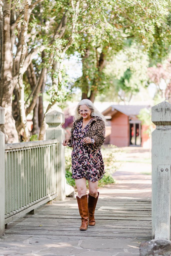smiling woman in a floral dress and leopard print jacket with brown cowboy boots jogging along a wooden bridge