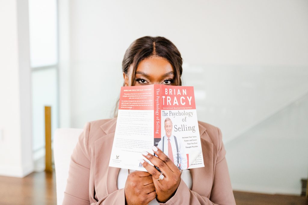 woman holding a book up and hiding part of her face. the book is called "The Psychology of Selling" by Brian Tracy