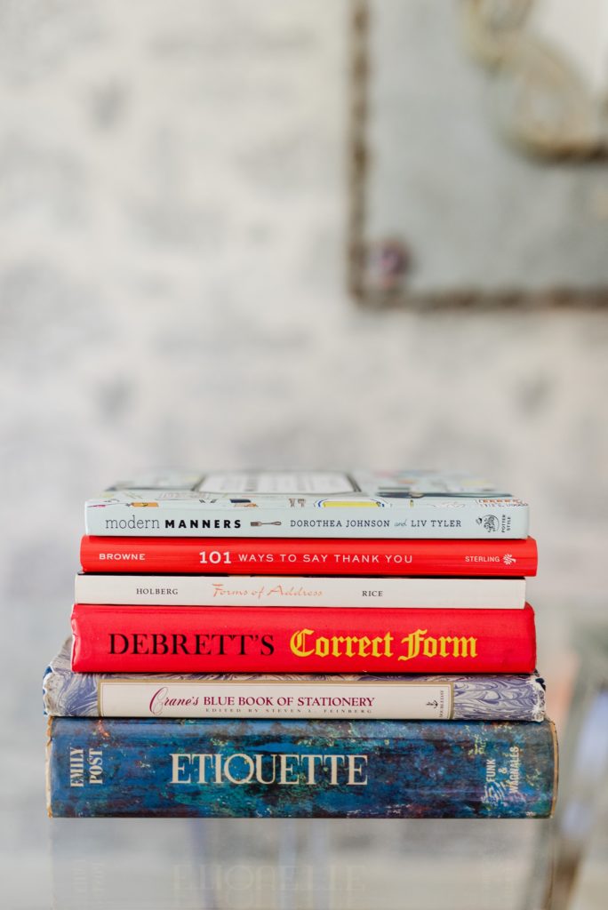 stack of books on a clear glass surface. book titles: modern manners by Dorothea Johnson and Liv Tyler, 101 Ways to Say Thank You, Forms of Address by Holberg and Rice, Debrett's Correct Form, Crane's Blue Book of Stationery, and Etiquette by Emily Post