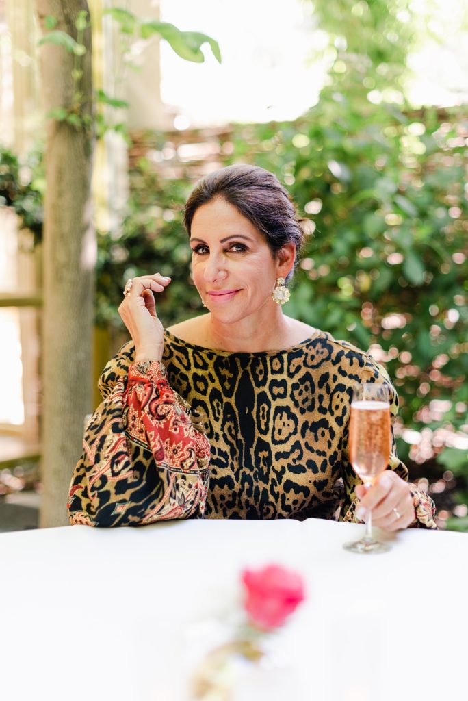 woman wearing animal print blouse is holding a champagne flute, her free hand is lifted by the side of her face with her elbow resting on the white table in front of her