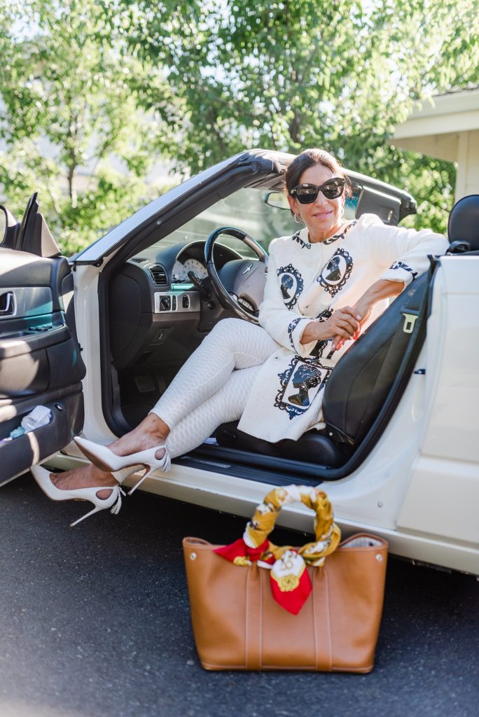 woman in white wearing shades is sitting on the driver's seat of a convertible with it's roof and door open. her brown leather handbag is on the ground next to the car