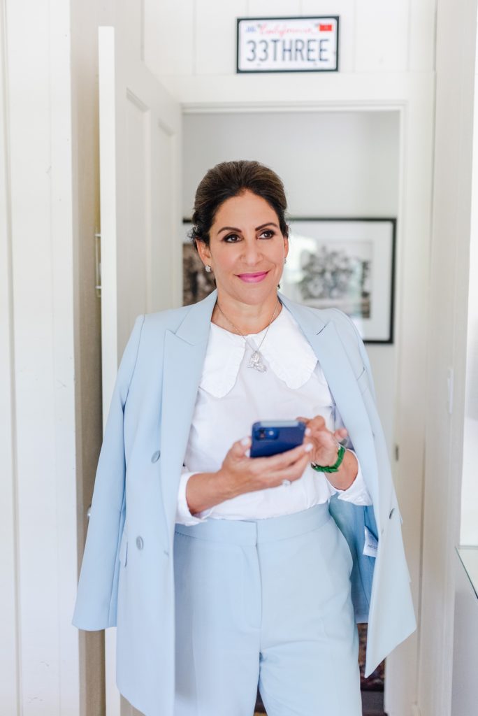 woman in white blouse and blue suit is holding her phone while standing by a doorway