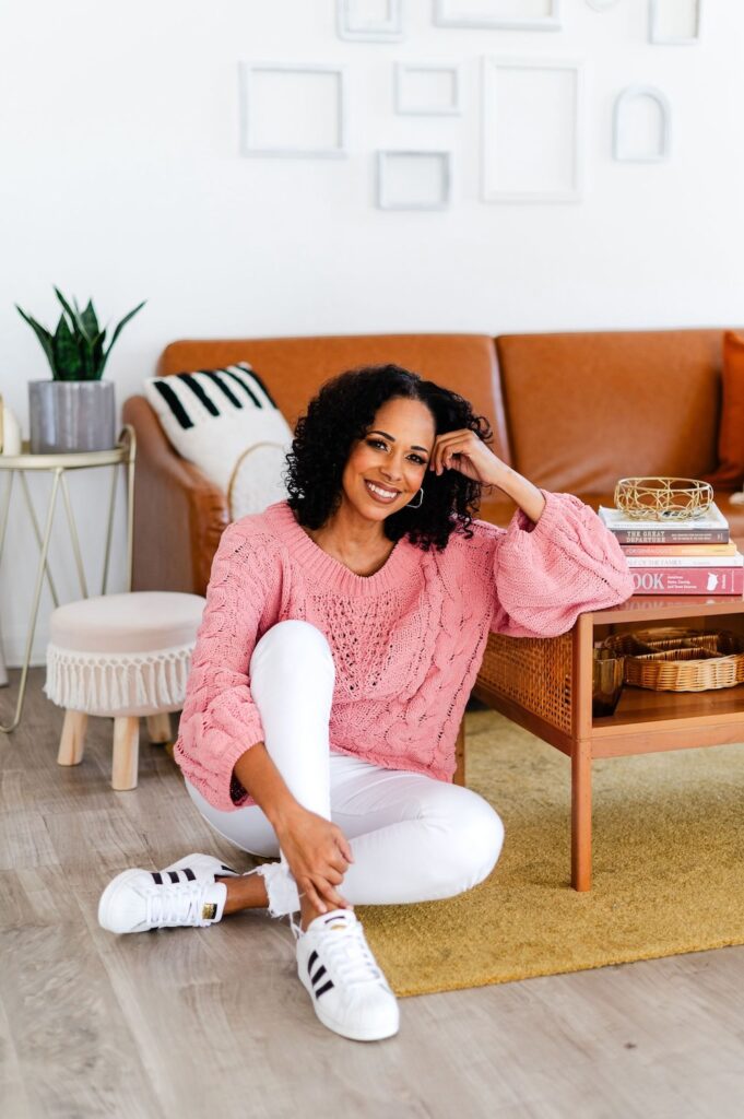 portrait of a professional genealogist wearing a pink sweater and white pants, sitting on the floor leaning on a coffee table