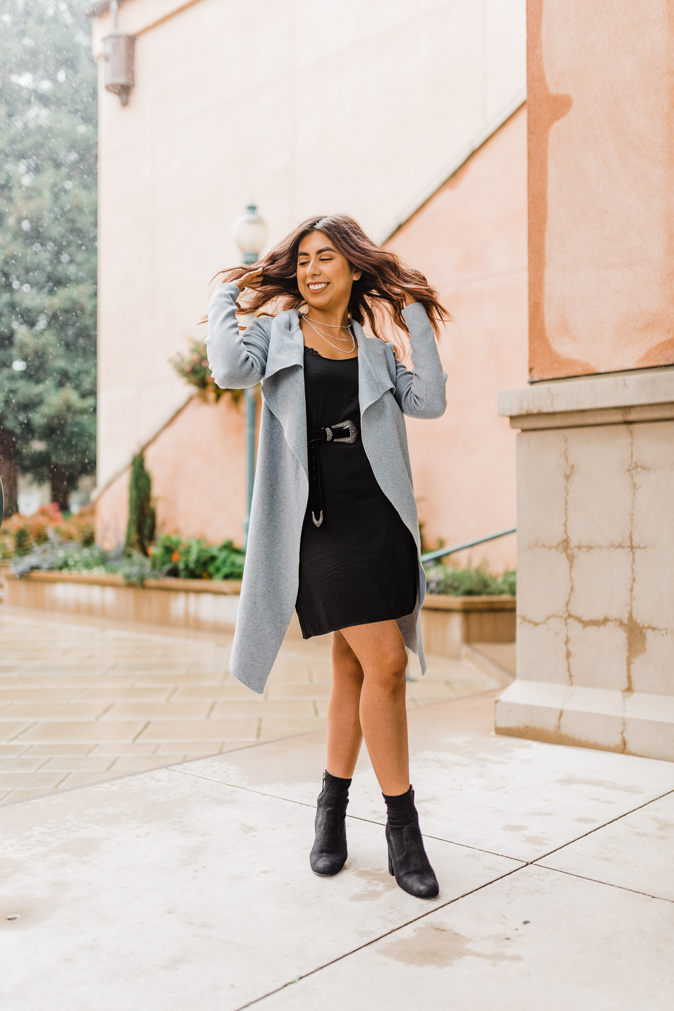 Fashion blogger doing a hair flip. Wearing a black dress with a long gray sweater.