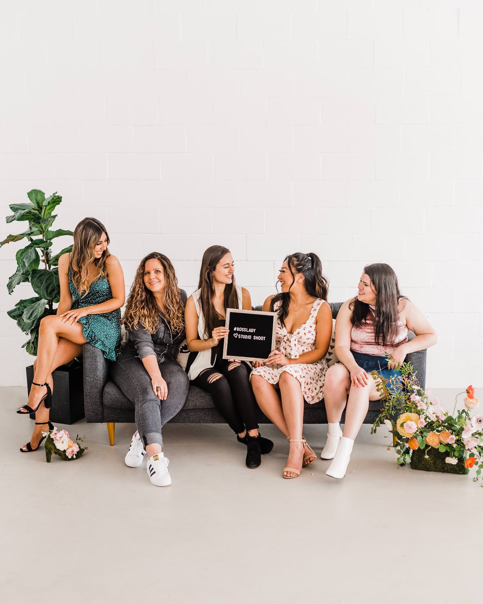 5 women sitting on a couch with plants on both sides. Two women in the middle are holding a sign that says "#BossLady studio shoot."