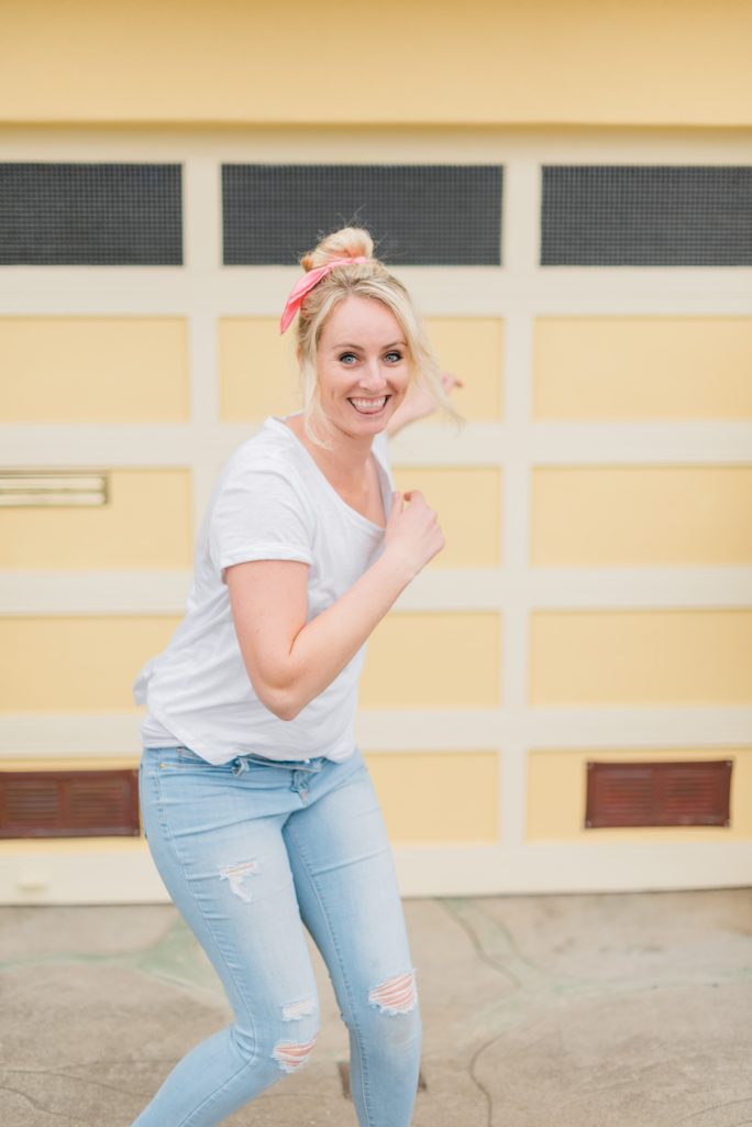 woman wearing white shirt, light denim jeans, and pink scrunchie in a fun jumping pose