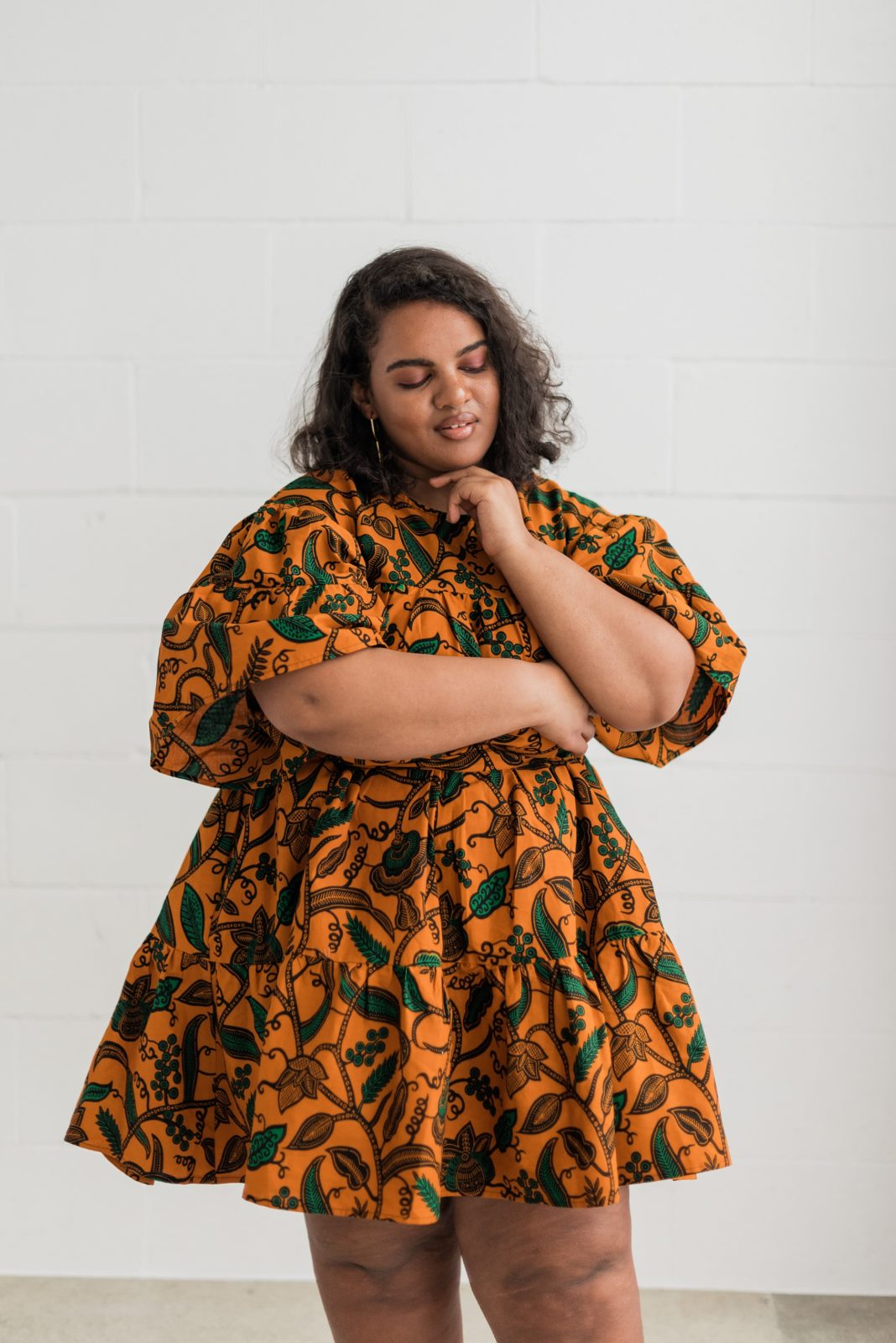 A black woman wearing a flared orange, patterned dress. She's leaning her chin on her arm.