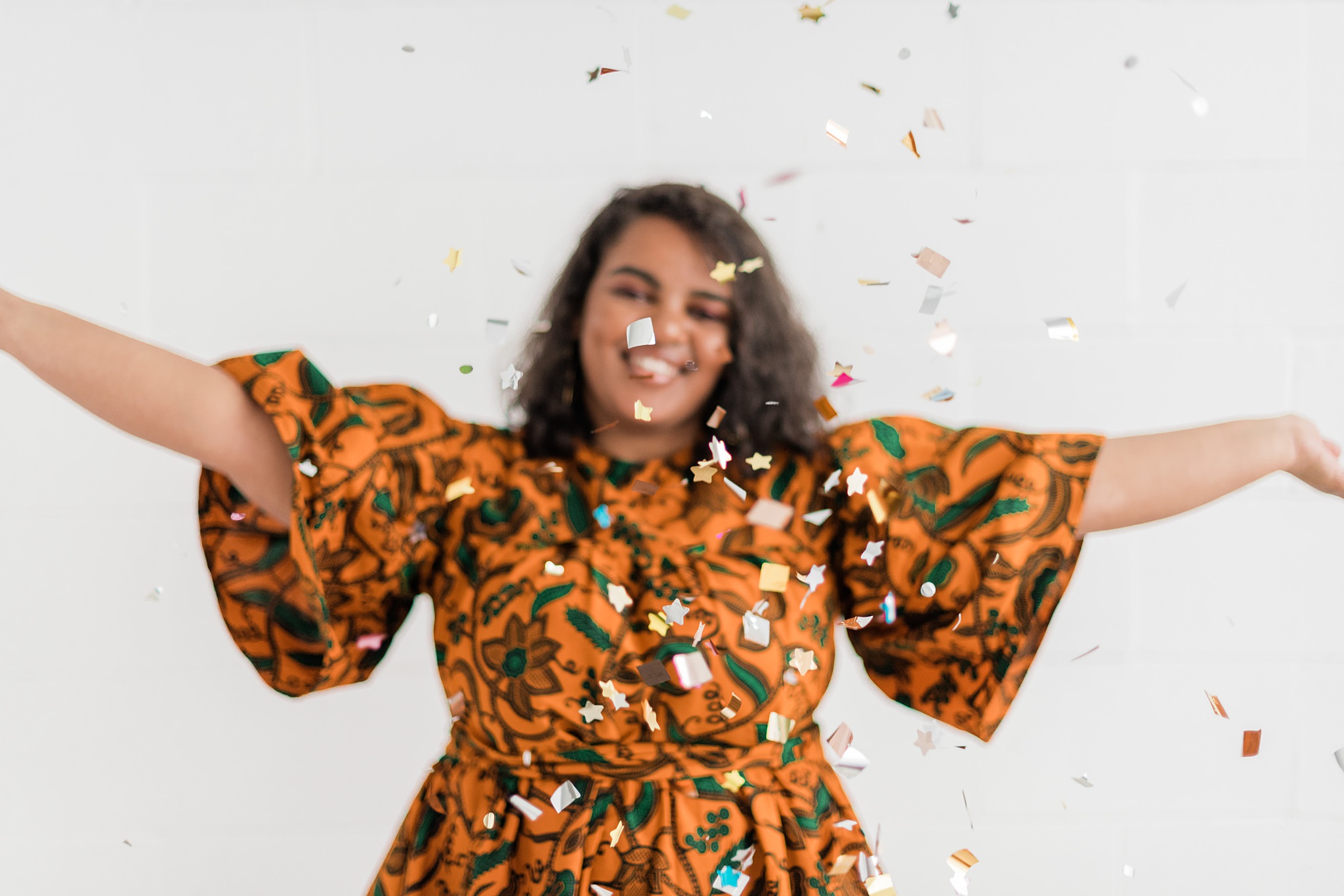 A black woman wearing a flared orange, patterned dress. The focus is on the confetti she's throwing.
