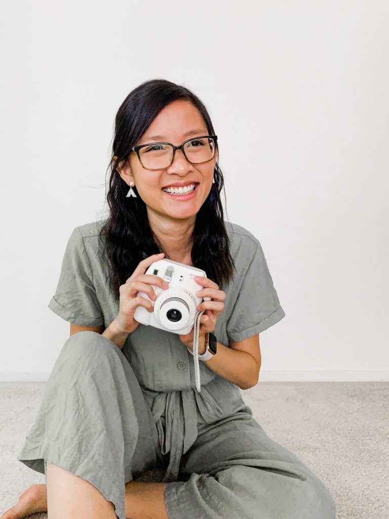 Woman wearing a green jumpsuit, sitting on the floor holding a fujifilm Instax camera.