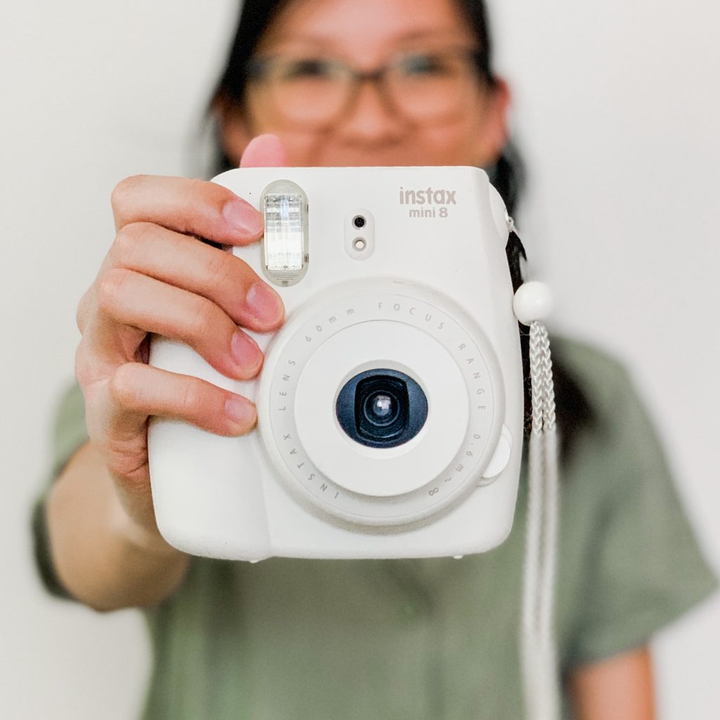 Woman wearing a green jumpsuit, sitting on the floor holding a fujifilm Instax camera.