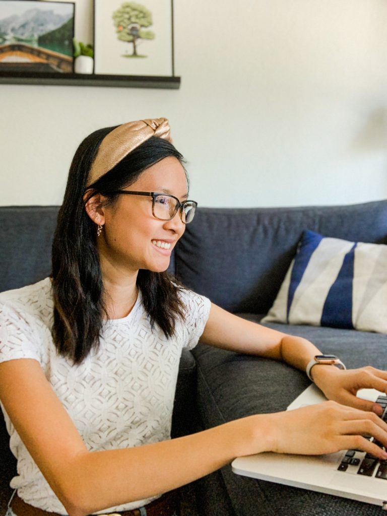 Woman wearing a white shirt and gold headband sitting on the floor. Her laptop is on the couch.