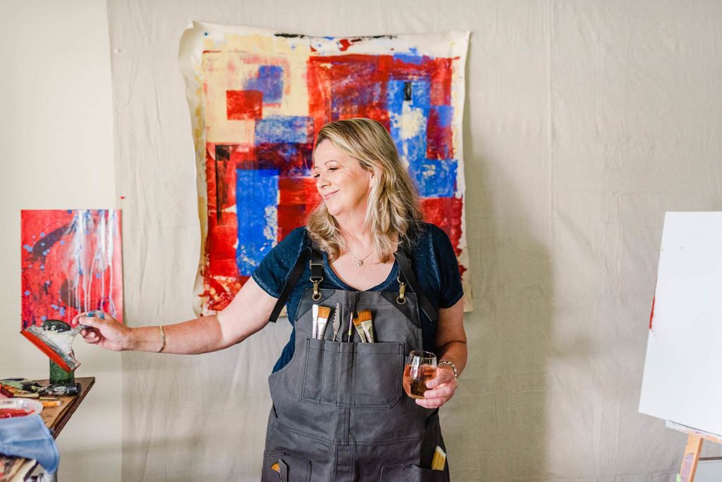 Portrait of an artist in her studio. She's wearing an apron with paint brushes and is holding a glass of wine.