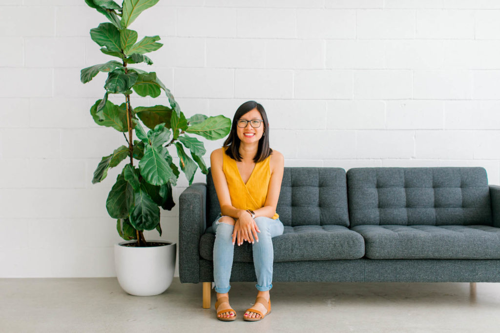 woman wearing a yellow shirt sitting on a gray couch. there's a fiddle leaf fig by the couch.