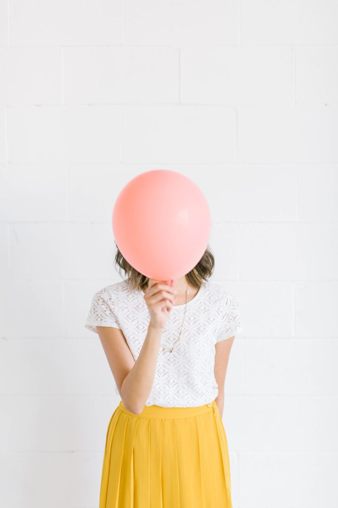 Woman holding a pink balloon in front of her face. She's wearing a white lace shirt and yellow skirt against a white wall.