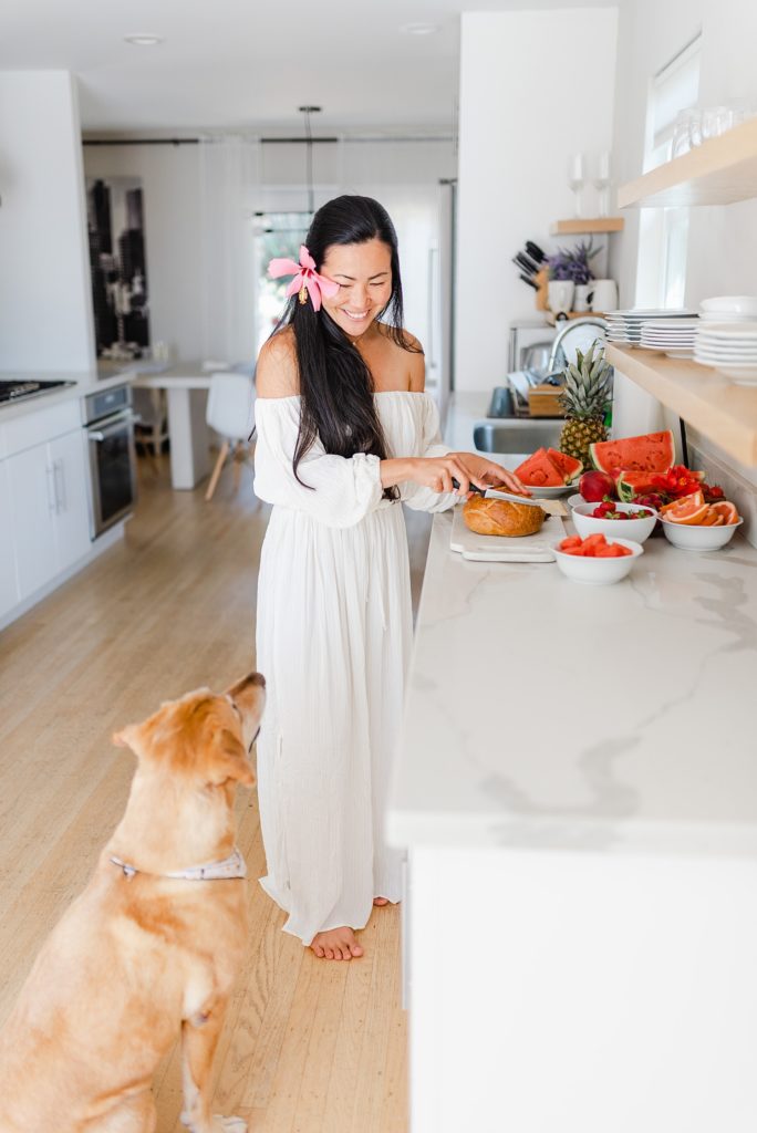 Woman in a long white dress standing by the kitchen counter while slicing bread. She is looking down at the brown dog beside her.