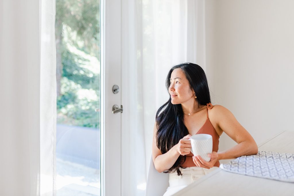 Woman wearing brown tank top sitting down by a glass door while looking outside. She is holding a white mug.