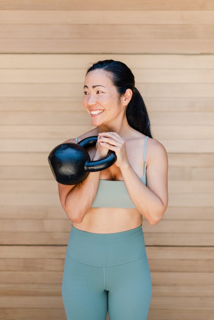 Woman in grey workout clothes is holding a kettlebell to her chest while her head is turned to the side.