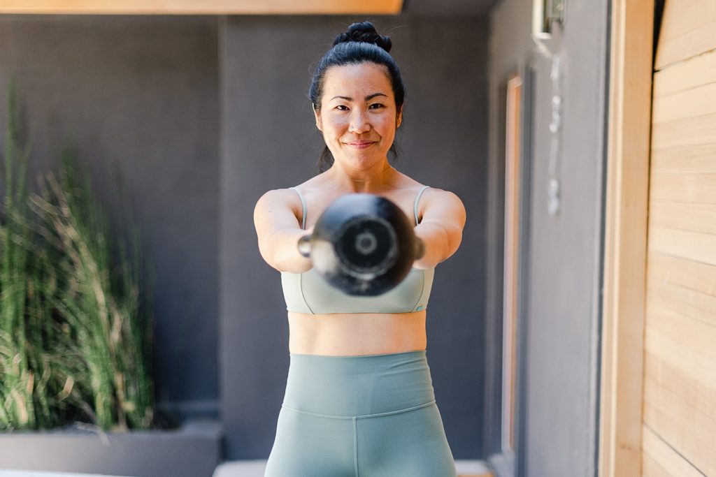 Woman in grey workout clothes is holding a kettle bell away from her body and pointed at the camera.