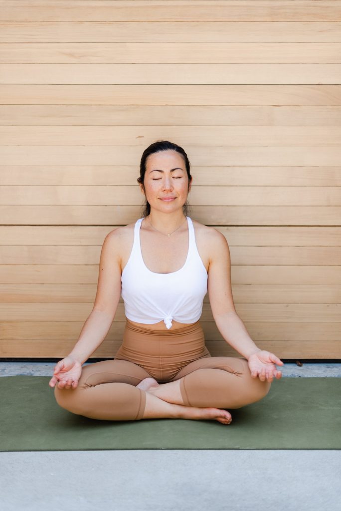 Woman in white tank top is sitting on the ground with her legs crossed. She is meditating.