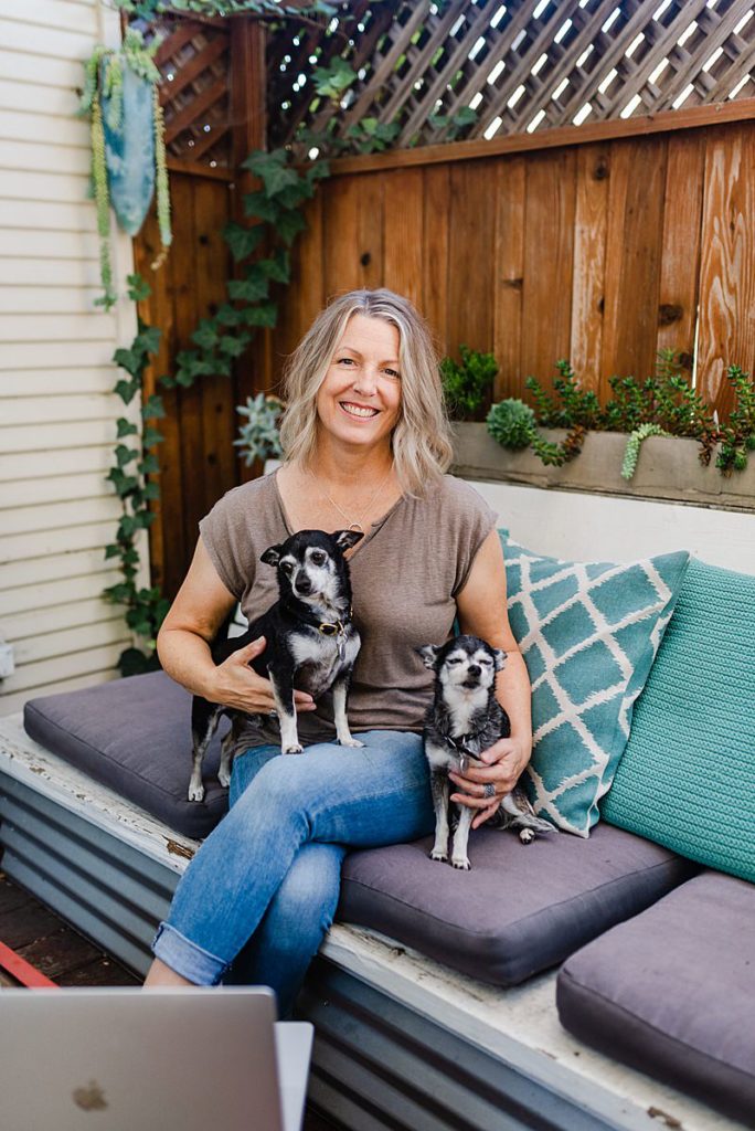 woman in brown blouse is sitting on a sofa in a patio of sorts with one small dog in her lap and another one beside her